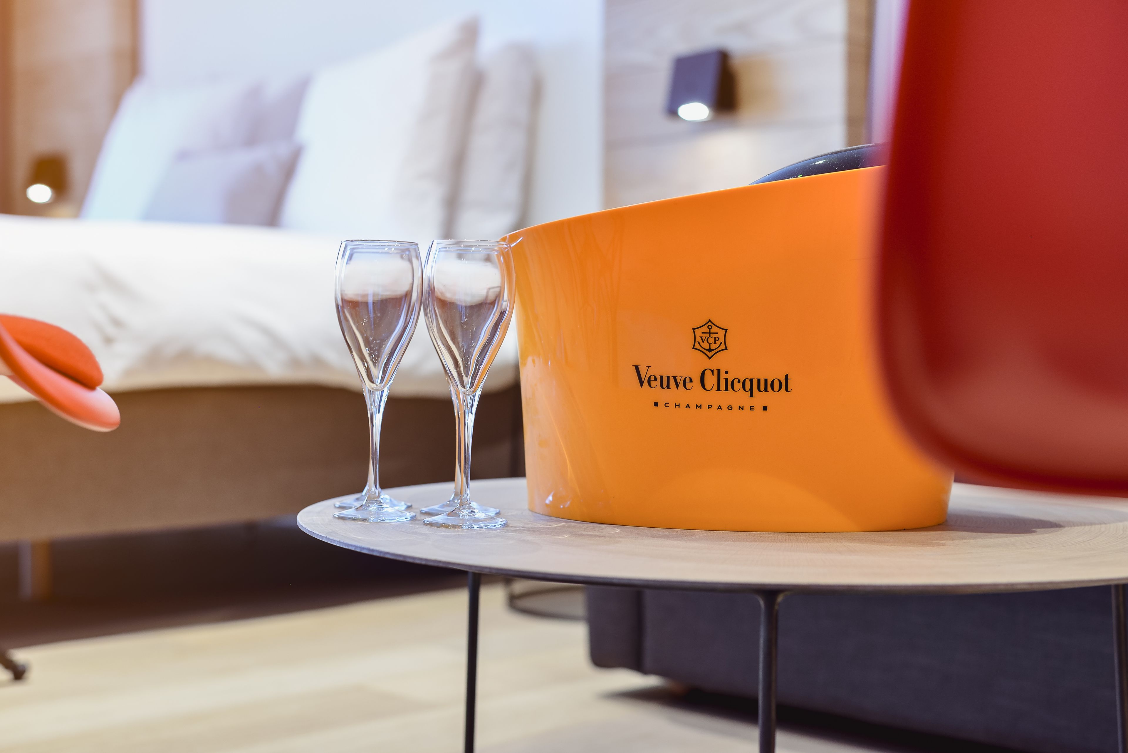 Ein Bett mit weißer Bettwäsche, ein Nachttisch mit einer Flasche Veuve Clicquot Champagner und zwei leere Gläser auf einem runden Tisch.