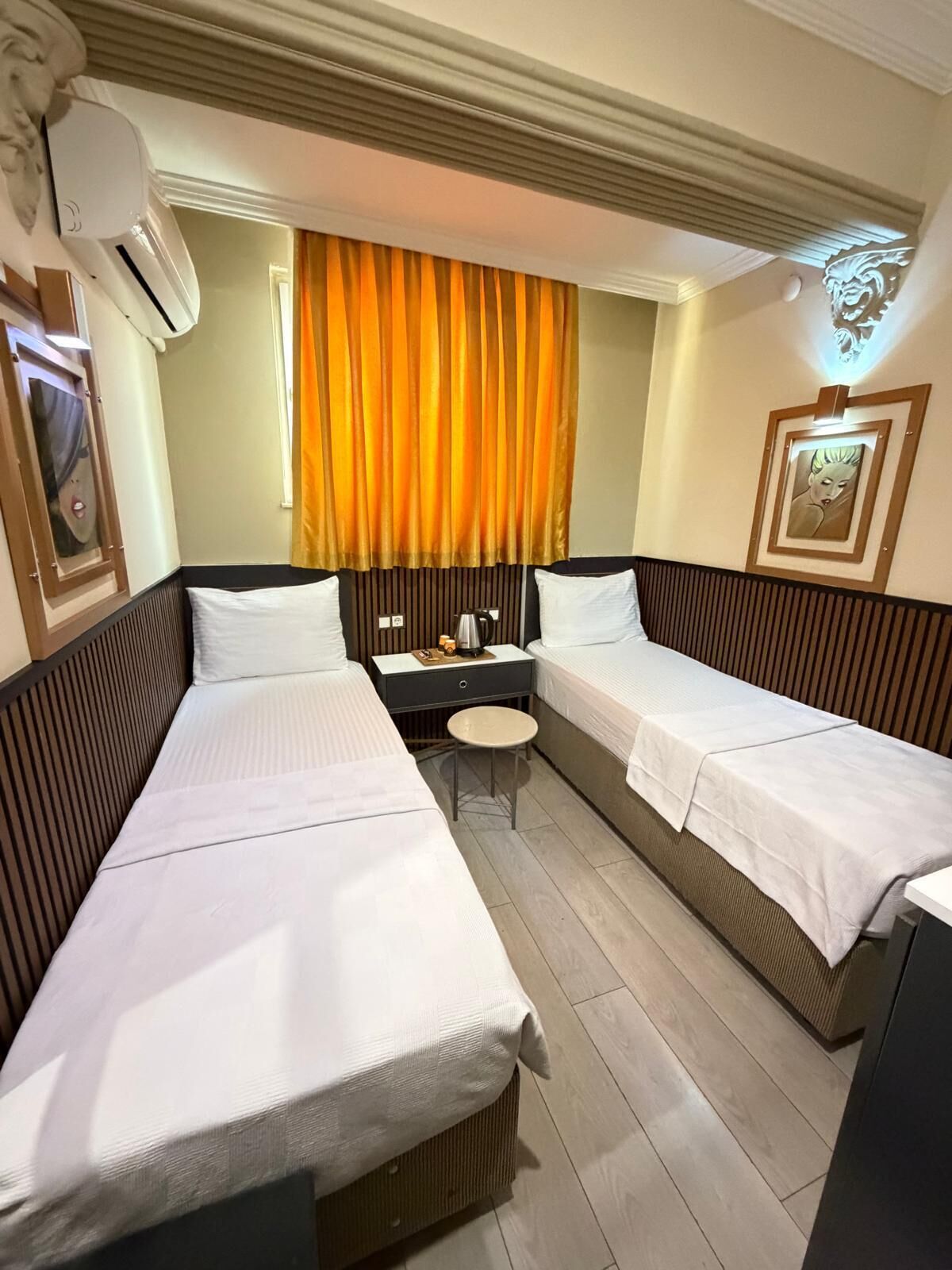 A hotel room with two single beds, a small table, a chair, an air conditioning unit, a window with curtains, and a wall-mounted light fixture.