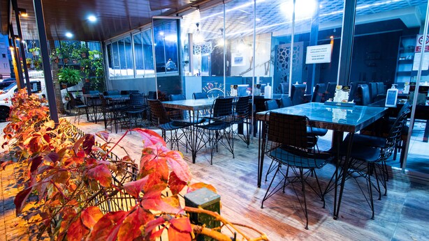 Un espacio de comedor al aire libre con mesas y sillas, rodeado de paredes de vidrio y plantas en macetas.