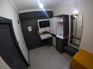 Une chambre d’hôtel de taille modeste, dotée d’une armoire de couleur sombre, d’un bureau avec une chaise, d’une télévision et d’un grand miroir.