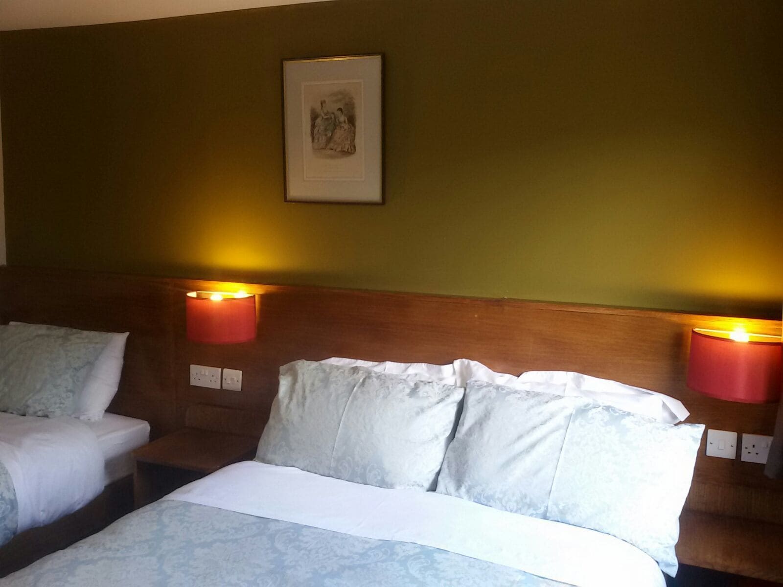 Quarto de hotel com duas camas, cada uma com lençóis brancos e lençóis com padrão azul claro. Duas luminárias fixadas na parede com abajures vermelhos estão em ambos os lados das camas. Um quadro emoldurado está pendurado na parede acima das camas.
