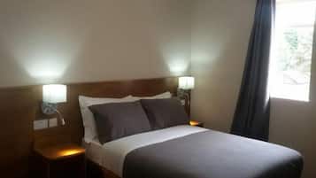 Habitación de hotel con cama, dos lámparas de noche, ventana con cortinas y mesita de noche con lámpara y toalla doblada.