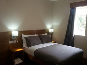 A hotel room with a bed, two bedside lamps, a window with curtains, and a nightstand with a lamp and a folded towel. - Crosshill House (Glasgow)