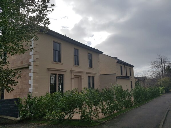 A two-story building with a beige facade, multiple windows, and a small garden area in front. - Crosshill House (Glasgow)