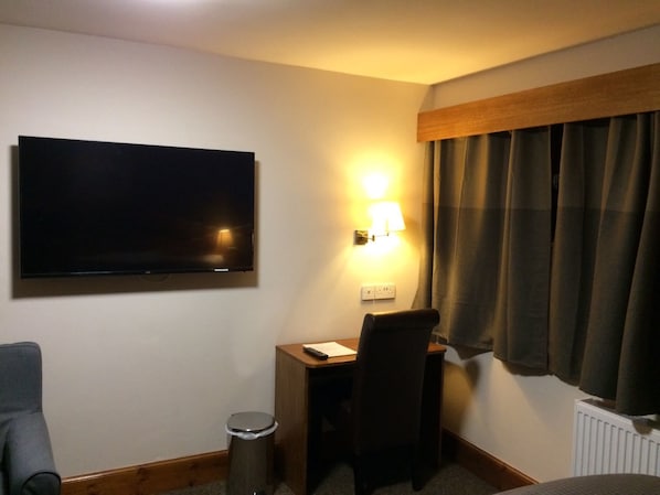 A hotel room with a flat-screen TV mounted on the wall, a desk with a chair, a small trash bin, and a window with curtains. - Crosshill House (Glasgow)