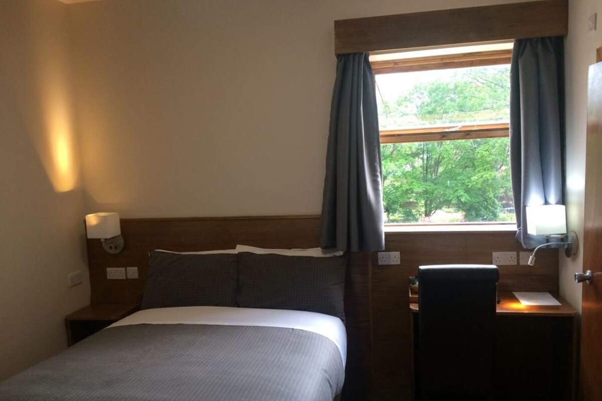 Una camera d'albergo con un letto, una scrivania, una sedia, una finestra con tende e un comodino con una lampada.