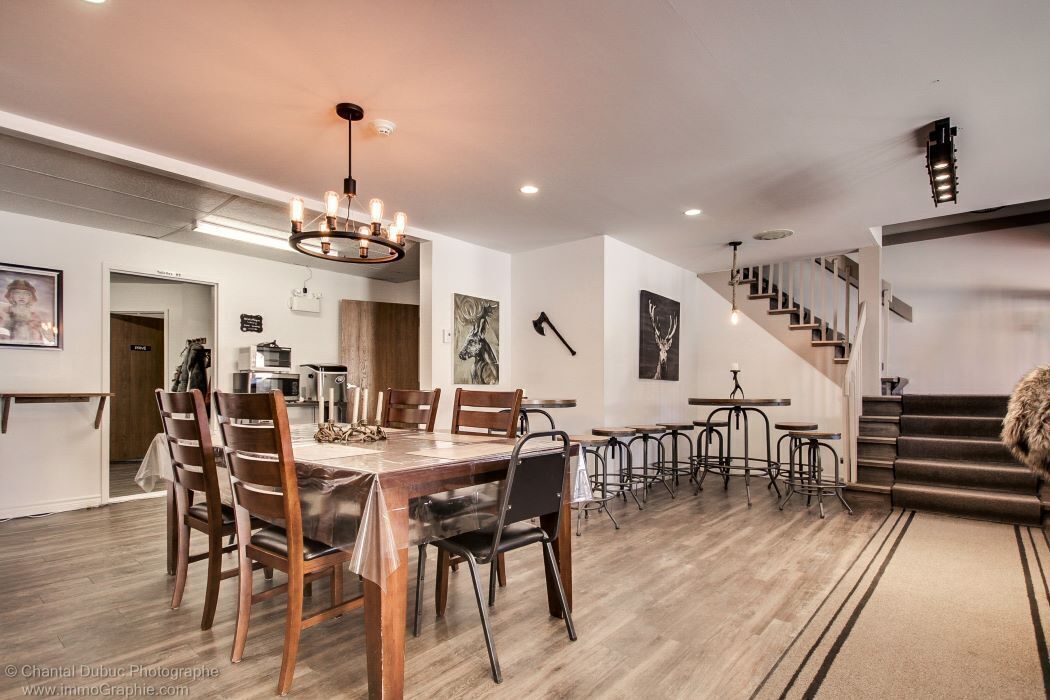 A spacious dining area with a long wooden table, multiple chairs, and a chandelier. There are bar stools along the wall and a staircase leading to another level.