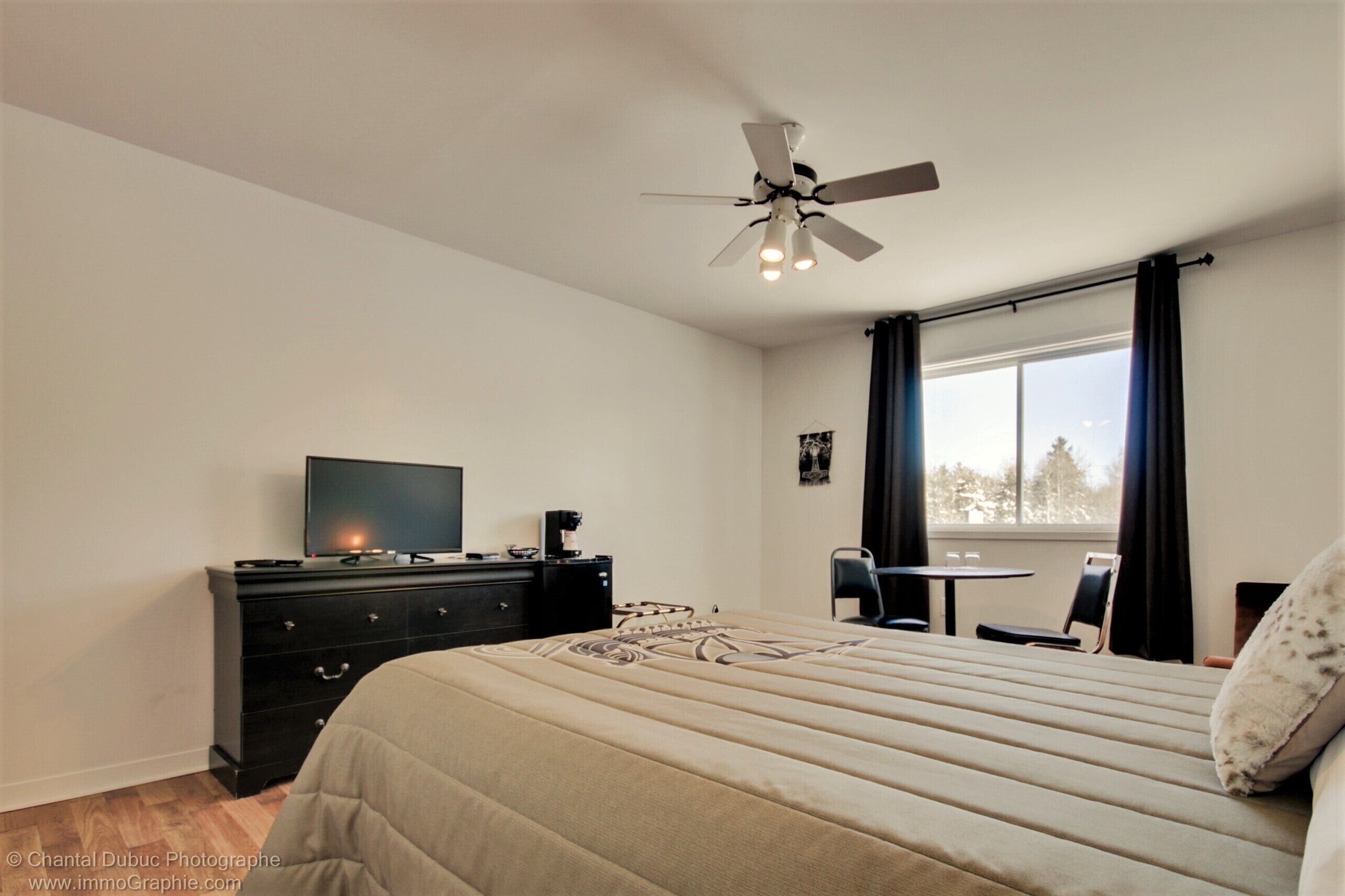 Un dormitorio con cama, televisor, cómoda, escritorio y ventana con cortinas.