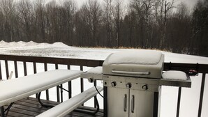 Ein Grill auf einer Holzterrasse mit Schnee auf dem Boden und umliegenden Bäumen.