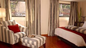 Una habitación de hotel con un sofá a rayas, un pequeño otoman, una cama con ropa de cama a rayas y cortinas.