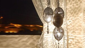 Un cuarto acogedor con un sofá estampado, una lámpara colgante y vistas a la puesta de sol a través de cortinas translúcidas.