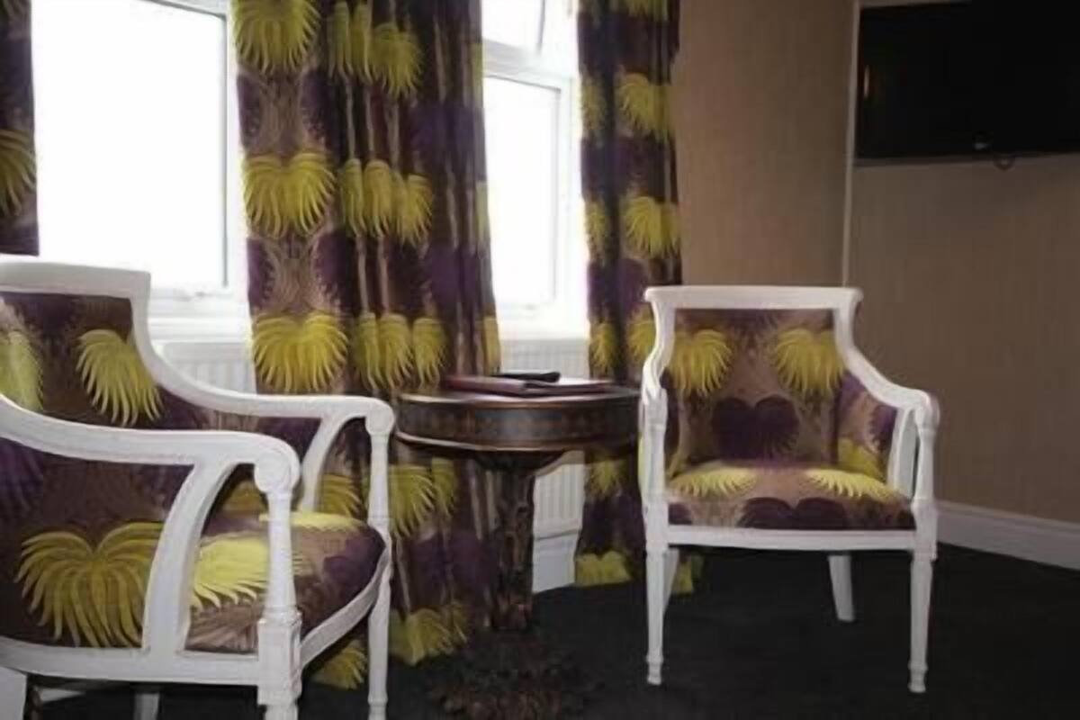 A room with two white armchairs and a small round table, a large window with curtains, and a flat-screen TV mounted on the wall.
