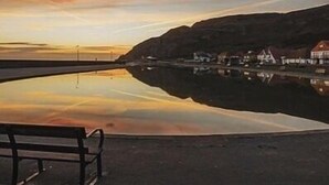 A bench overlooking a calm body of water with a reflection of a town and hills in the background. - The Lilly (Llandudno)