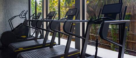 A row of treadmills with digital screens, positioned in a room with large windows overlooking a garden.