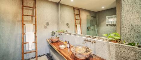 Un baño con tocador de madera, dos lavamanos de cerámica grandes, un espejo y zona de ducha.