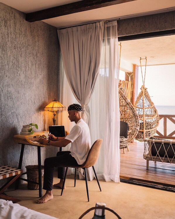 A room with a bed, a table with a laptop, a lamp, and a wicker chair.