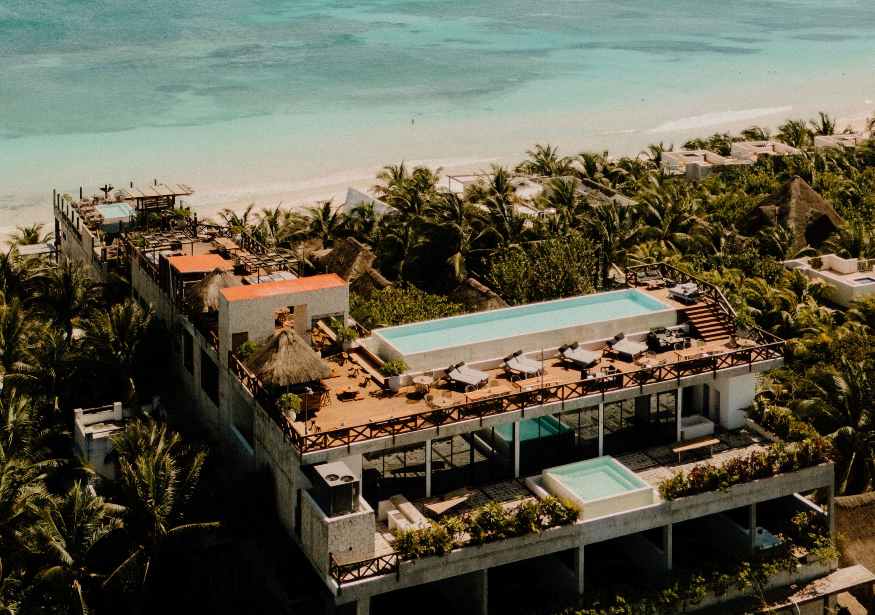 An aerial view of a resort with a large swimming pool, multiple lounge areas, and a beach view.