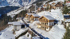 Ein verschneites Bergdorf mit Chalets, einer kurvenreichen Straße und schneebedeckten Berggipfeln im Hintergrund.