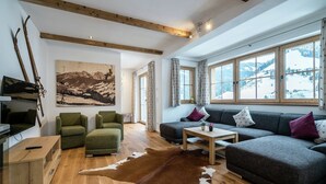 Ein gemütliches Wohnzimmer mit Holzbalkendecke, einem großen Fenster mit Blick auf Schnee, einem grünen Sofa, einem braunen Lederteppich und einem Gemälde von einer Berglandschaft.