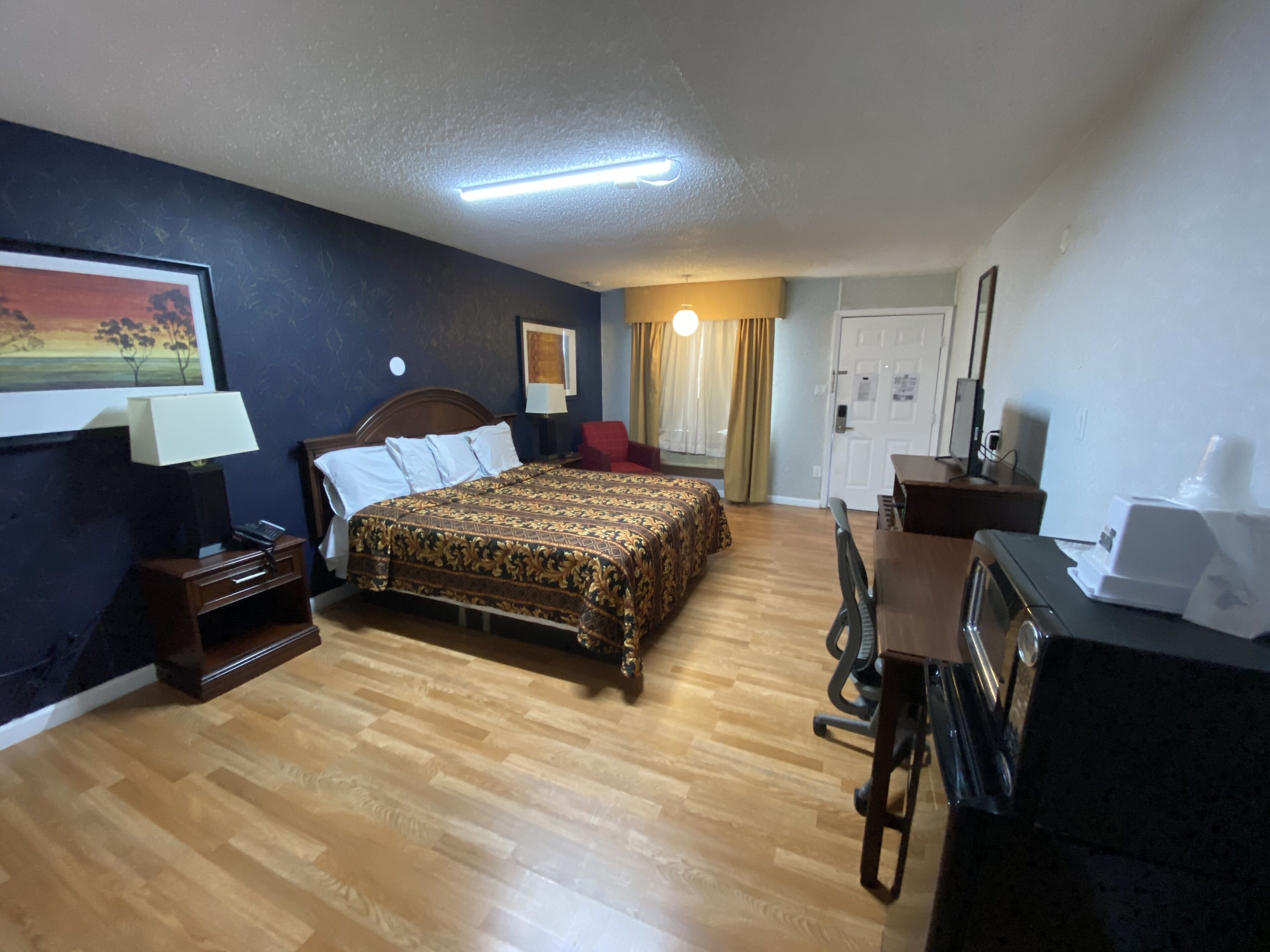 Habitación de hotel con piso de madera, una cama con colcha estampada, un escritorio con computadora, una silla, una lámpara y un cuadro en la pared.