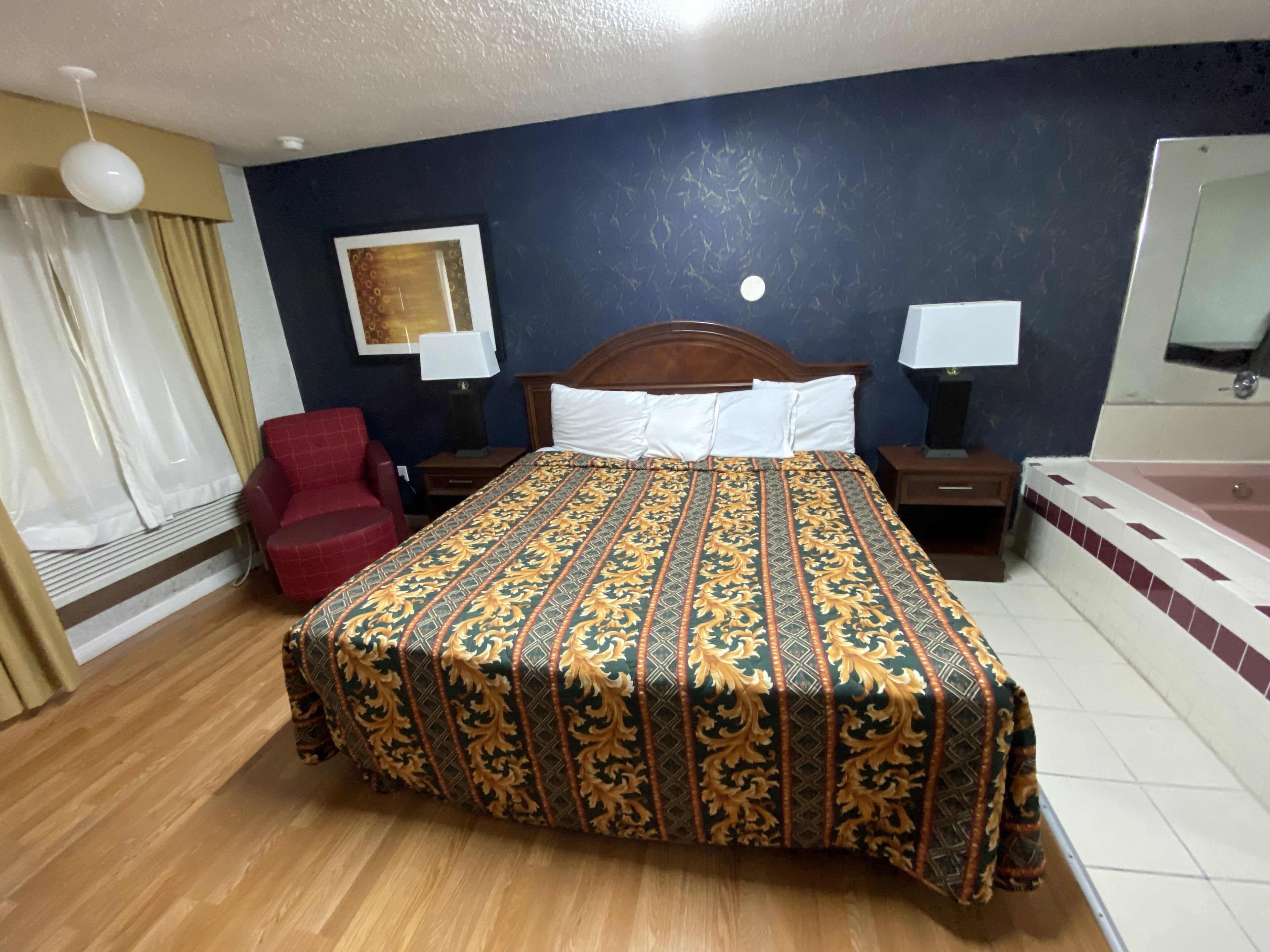Ein Hotelzimmer mit einem hölzernen Bett, einem roten Sessel, einem Nachttisch aus Holz, einem Beistelltisch und einem Badezimmer mit einer Badewanne.