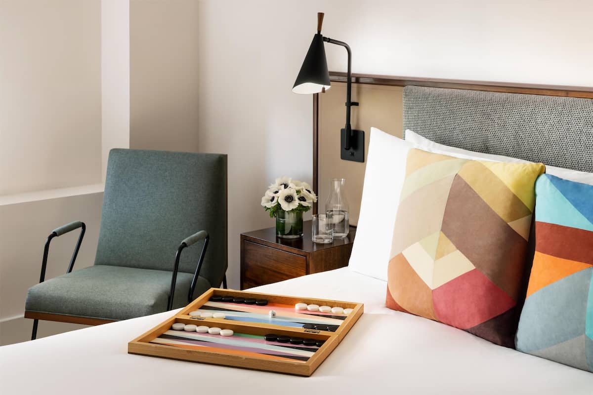 A hotel room with a bed, a chair, a bedside table with a lamp, and a decorative pillow.