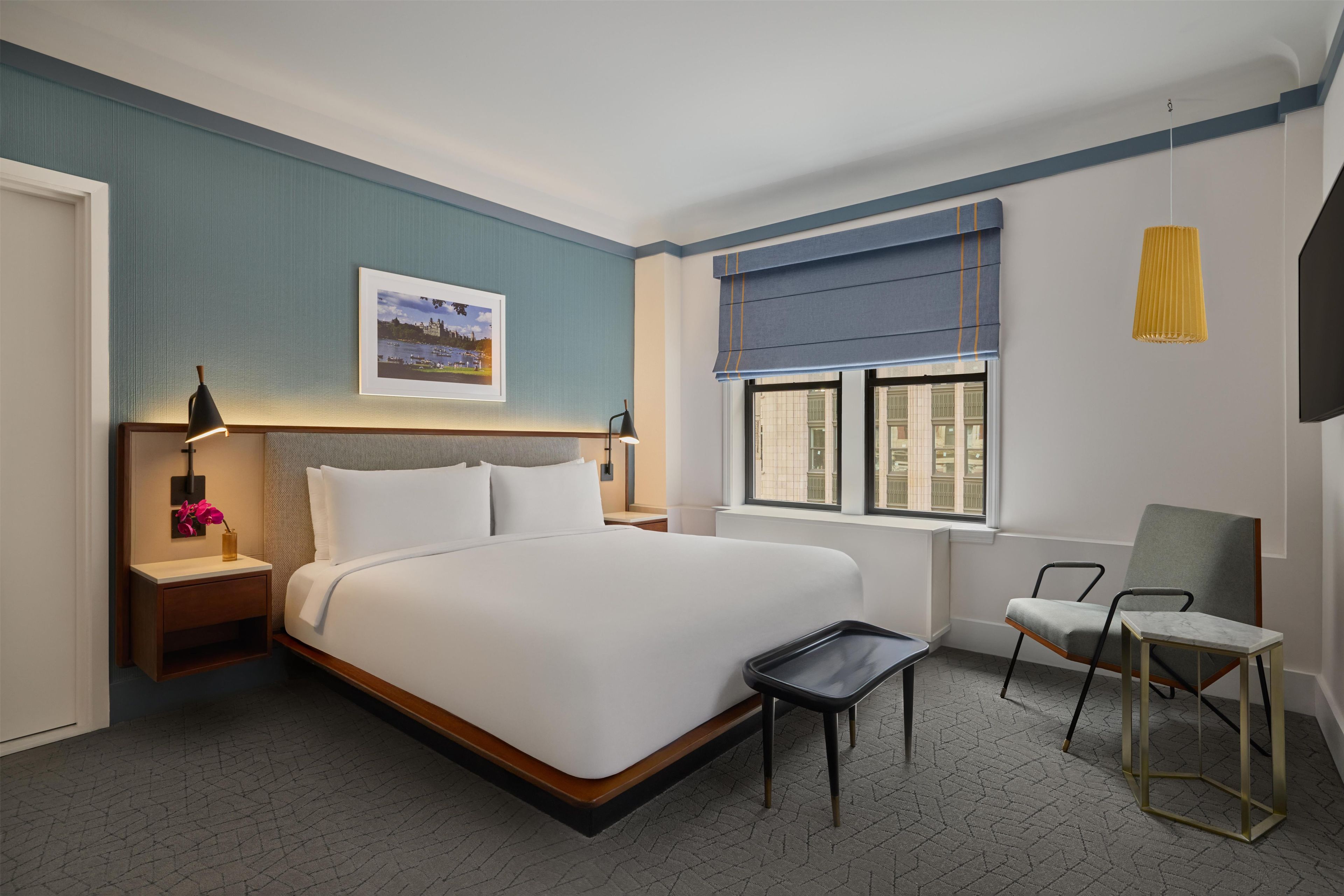A hotel room with a bed, bedside table, lamp, chair, window, and a picture on the wall.