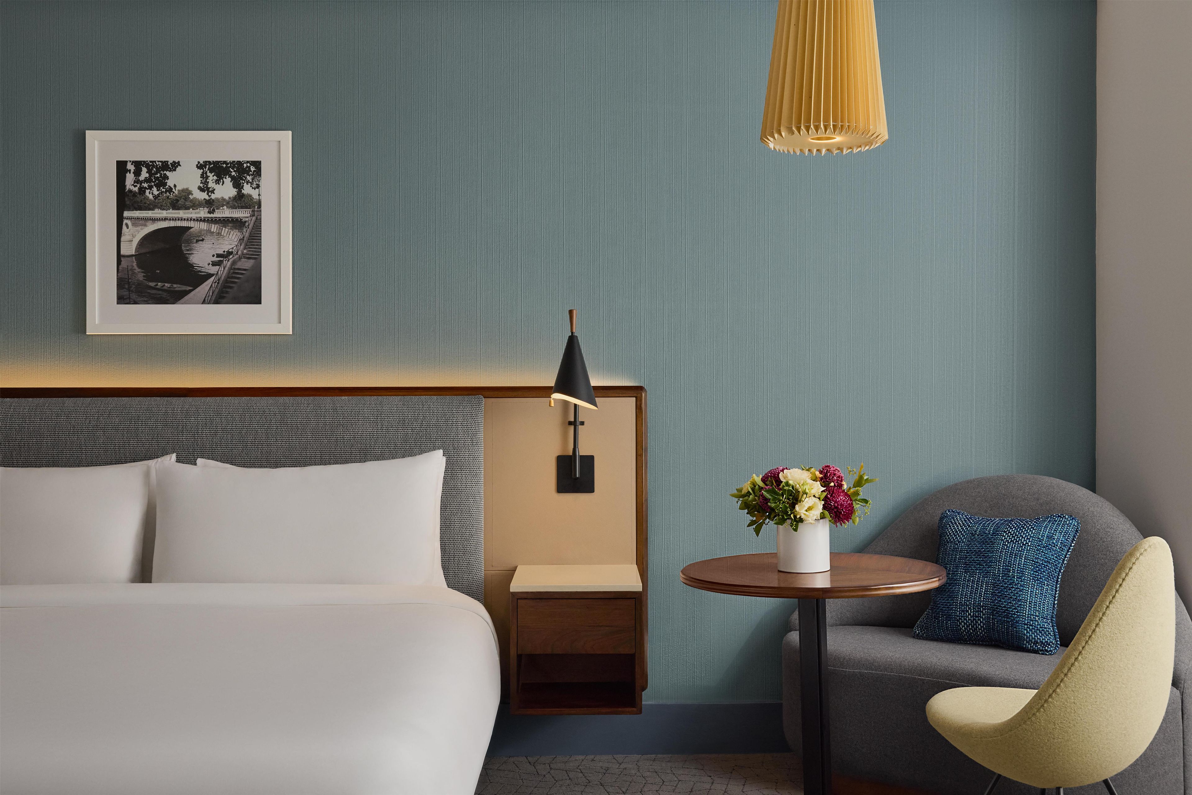 Une chambre d’hôtel avec un lit, une table de chevet, une petite table de nuit avec une lampe, une chaise et un tableau encadré au mur.