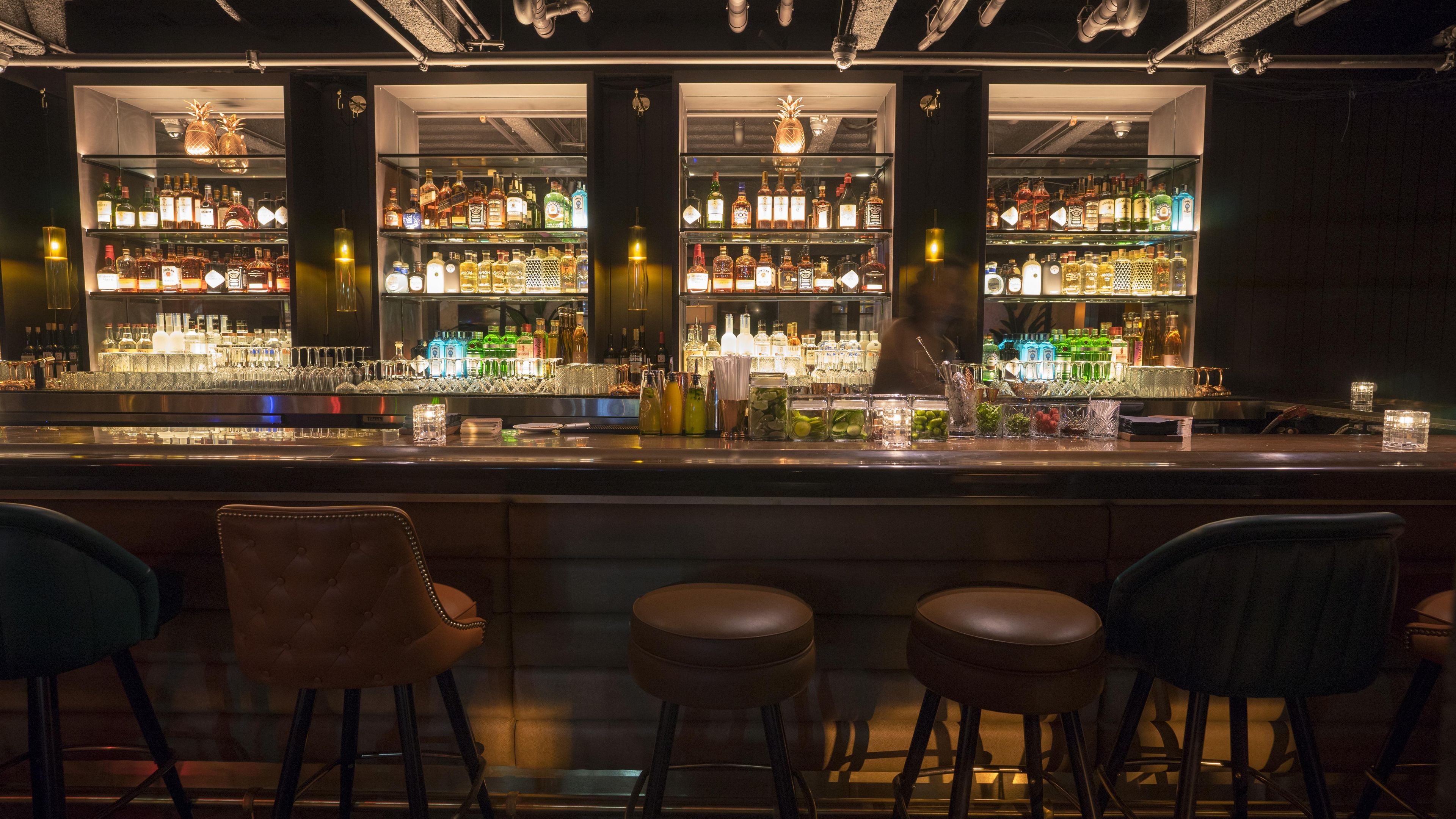 A bar area with multiple shelves of liquor bottles, a bar counter, and three leather bar stools.