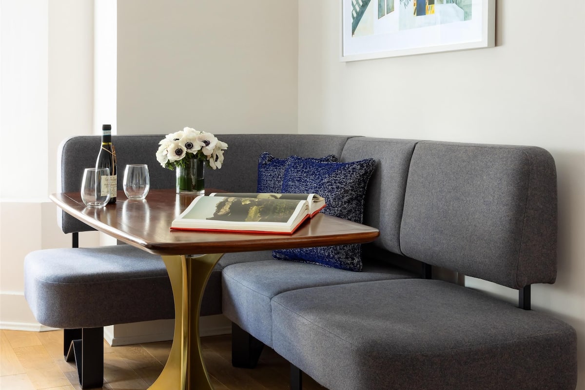 A modern living room with a grey sofa, a small wooden table with a book and glasses, a vase with flowers, and a hanging pendant light.