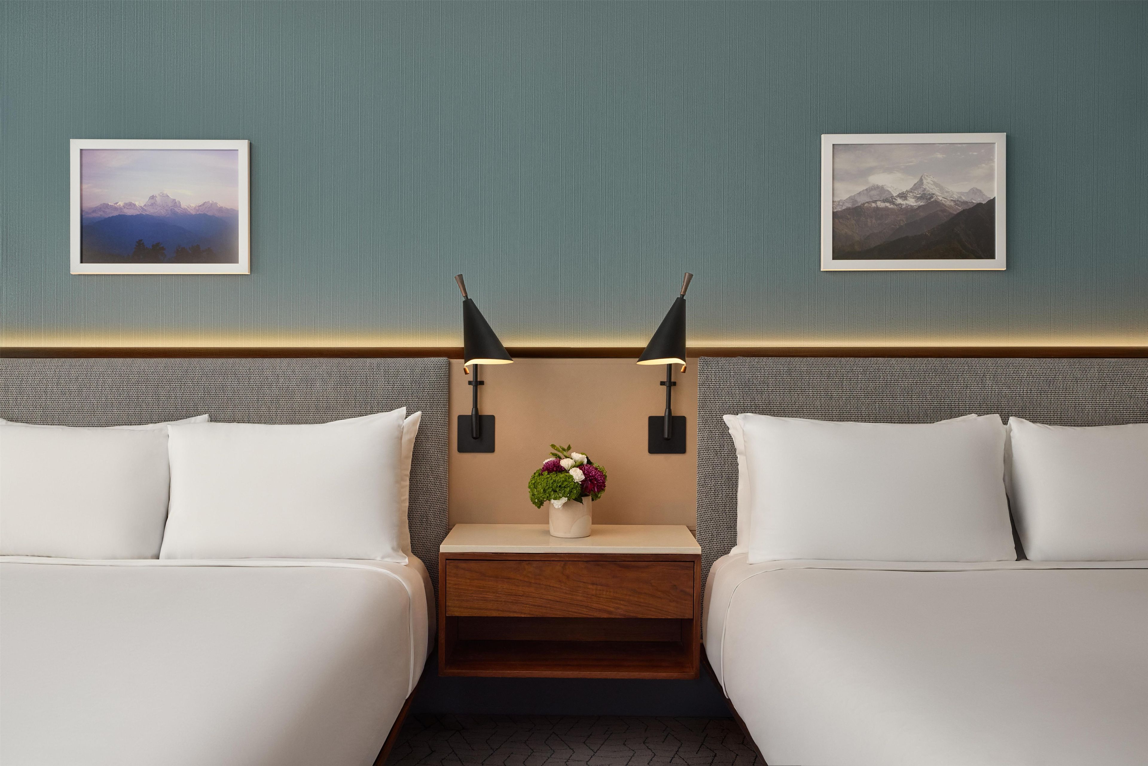 A hotel room with two beds, a bedside table with a vase of flowers, and two framed pictures on the wall.