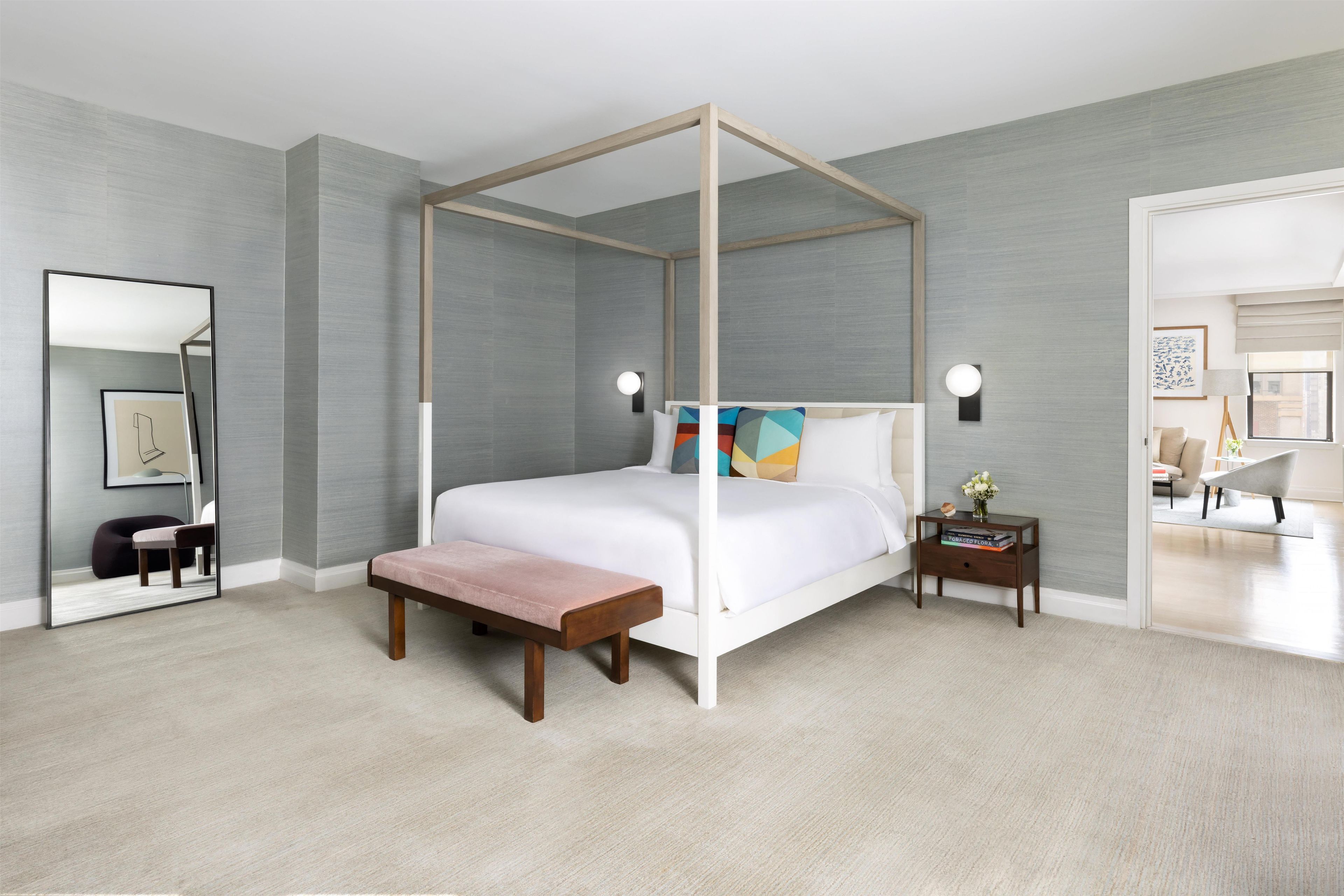 Een moderne slaapkamer met een hemelbed, een bankje, een nachtkastje met lamp en een spiegel.