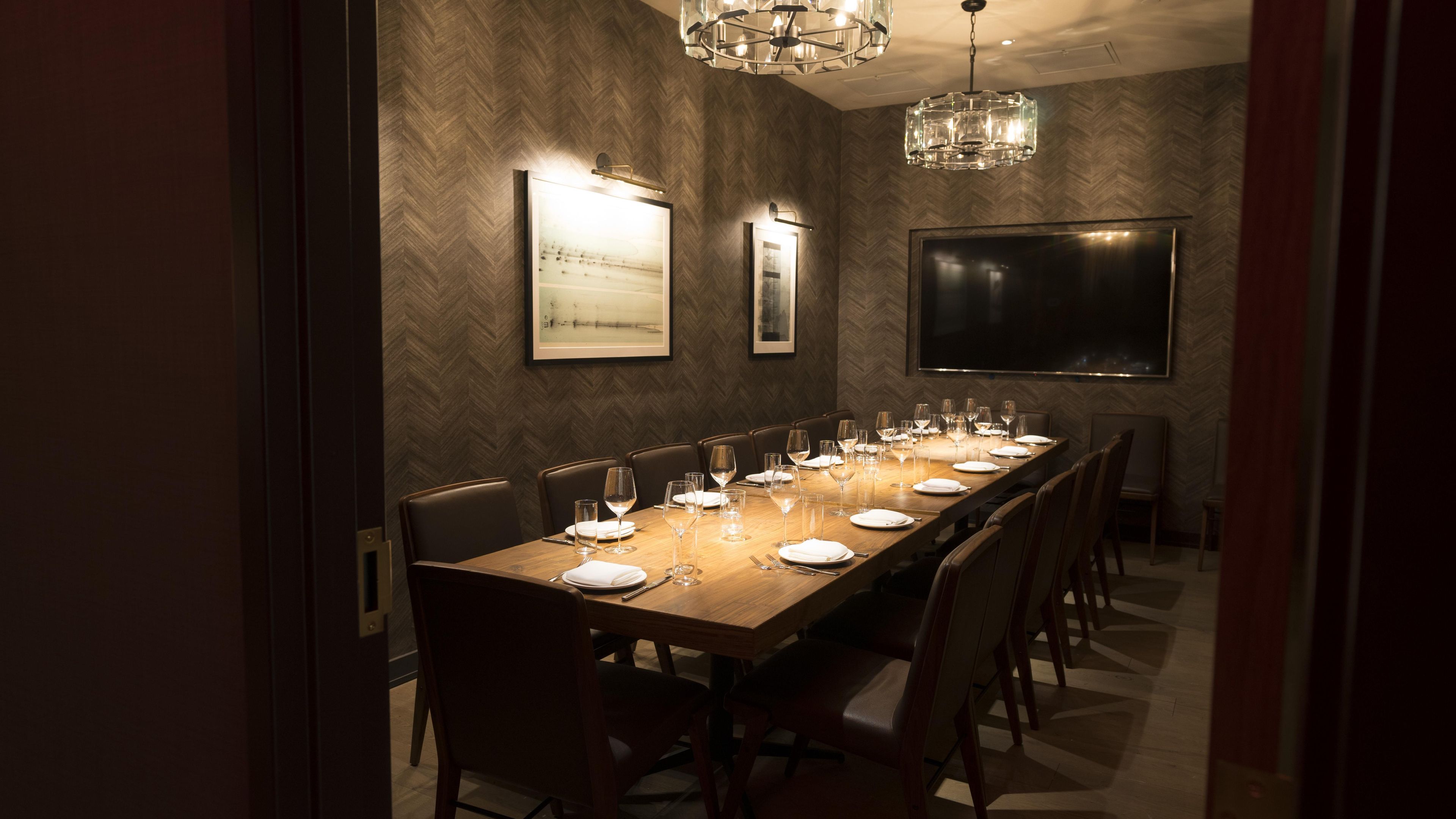 A long dining table set for a meal, flanked by chairs, in a room with a herringbone-patterned wall and a large flat-screen TV.