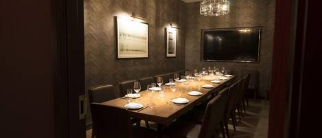 A long dining table set for a meal, flanked by chairs, in a room with a herringbone-patterned wall and a large flat-screen TV.