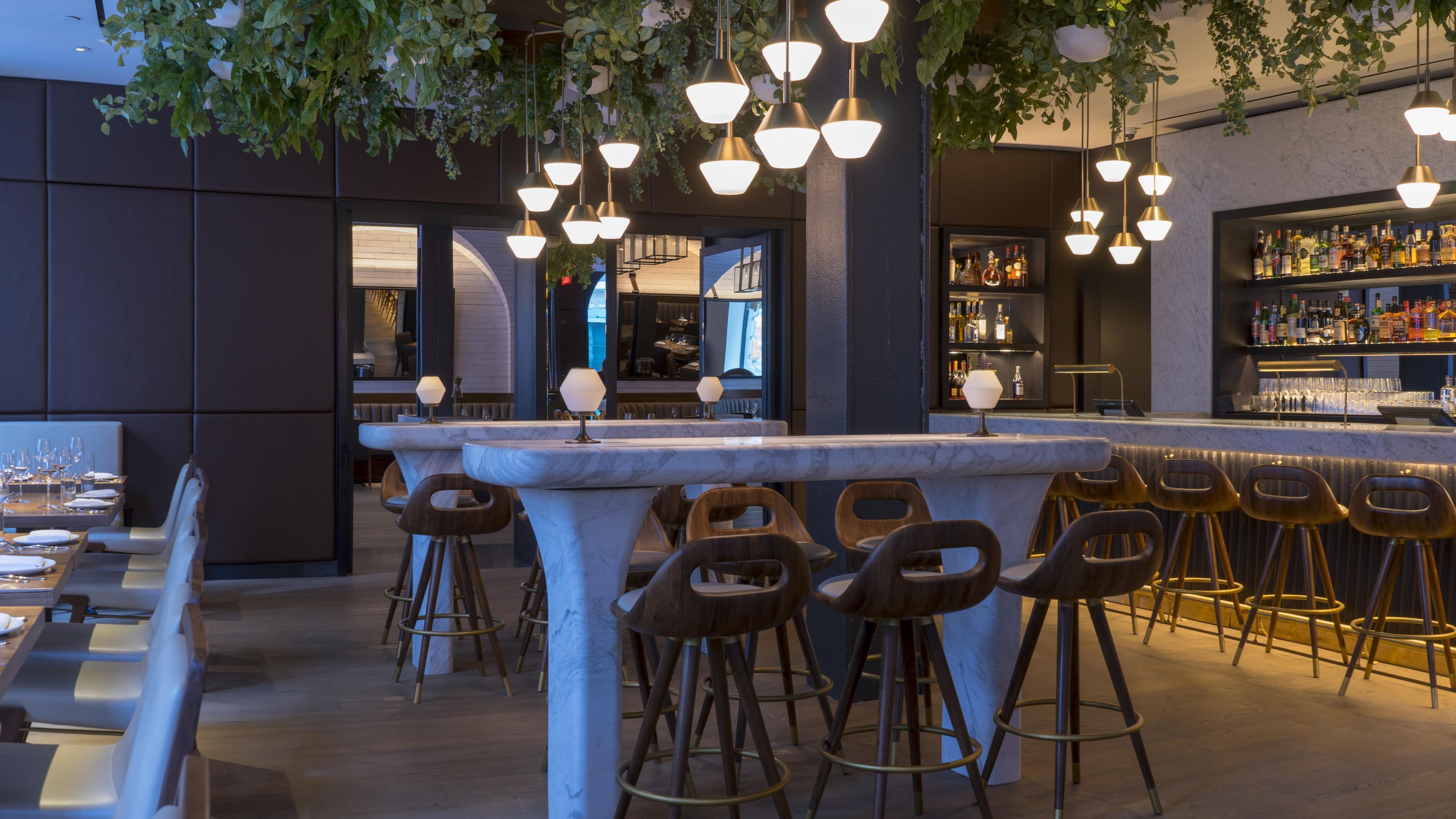 Een bar met een marmeren toonbank, houten krukken, hangende pendelverlichting en een bar gevuld met flessen.