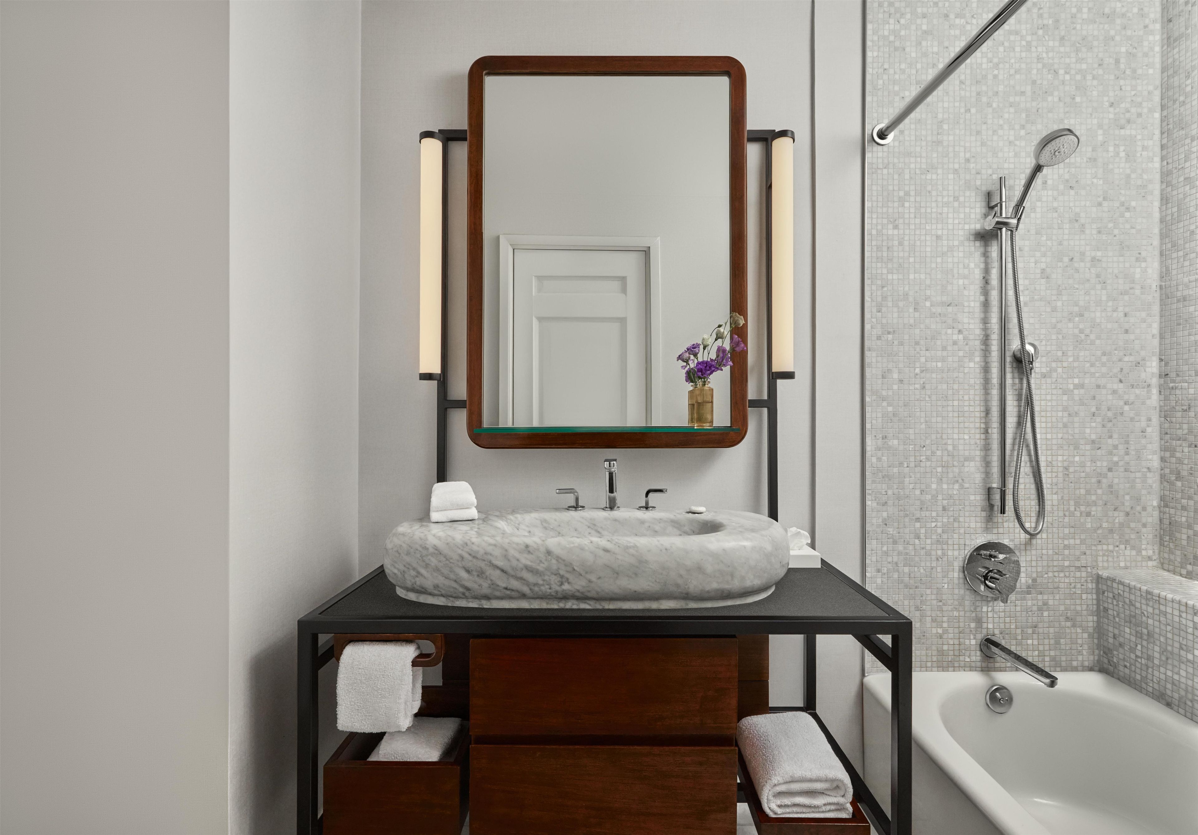A bathroom with a marble sink, a wooden vanity, a large mirror, a shower with a handheld showerhead, and a white bathtub.