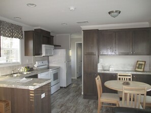 A modern kitchen with dark wood cabinets, a white refrigerator, a microwave, and a stove. There is a dining area with a wooden table and chairs. - Settler's Rest RV Resort (Zephyrhills)