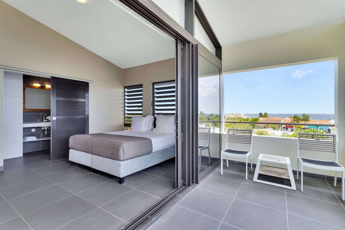 A modern bedroom with a bed, a small bathroom, and a balcony with chairs and a table.