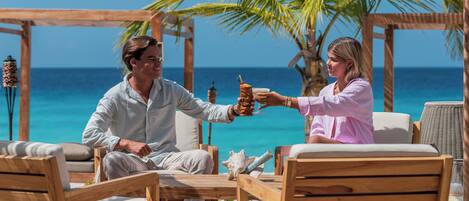 A wooden deck with two people seated at a table, a palm tree, and a view of the ocean.