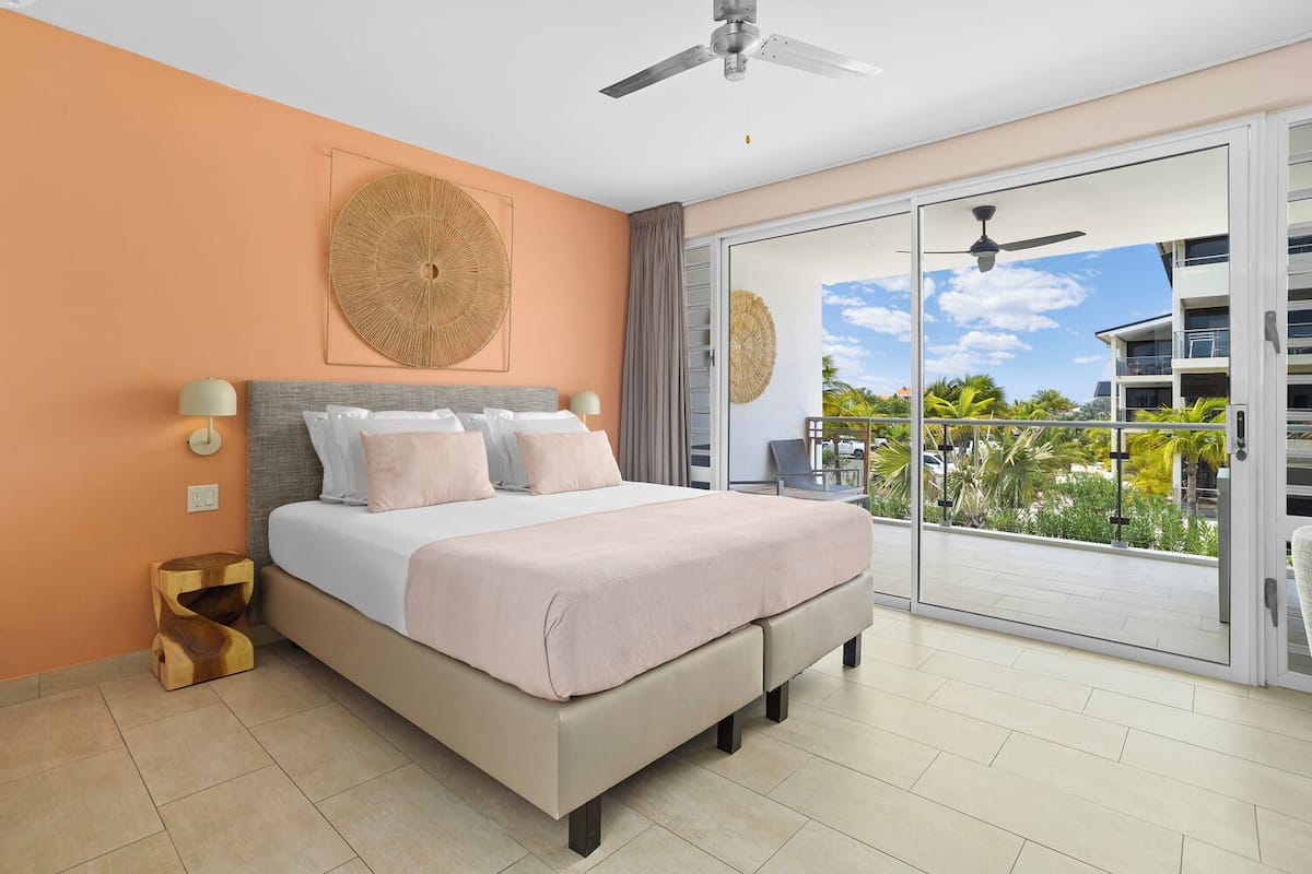 A bedroom with a bed, a ceiling fan, a large window with a view of a building and palm trees, and a balcony with a chair.