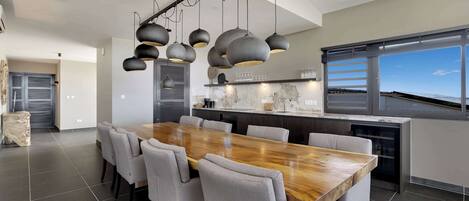 A modern kitchen with a wooden dining table and chairs, a large window, and a view of the sky.