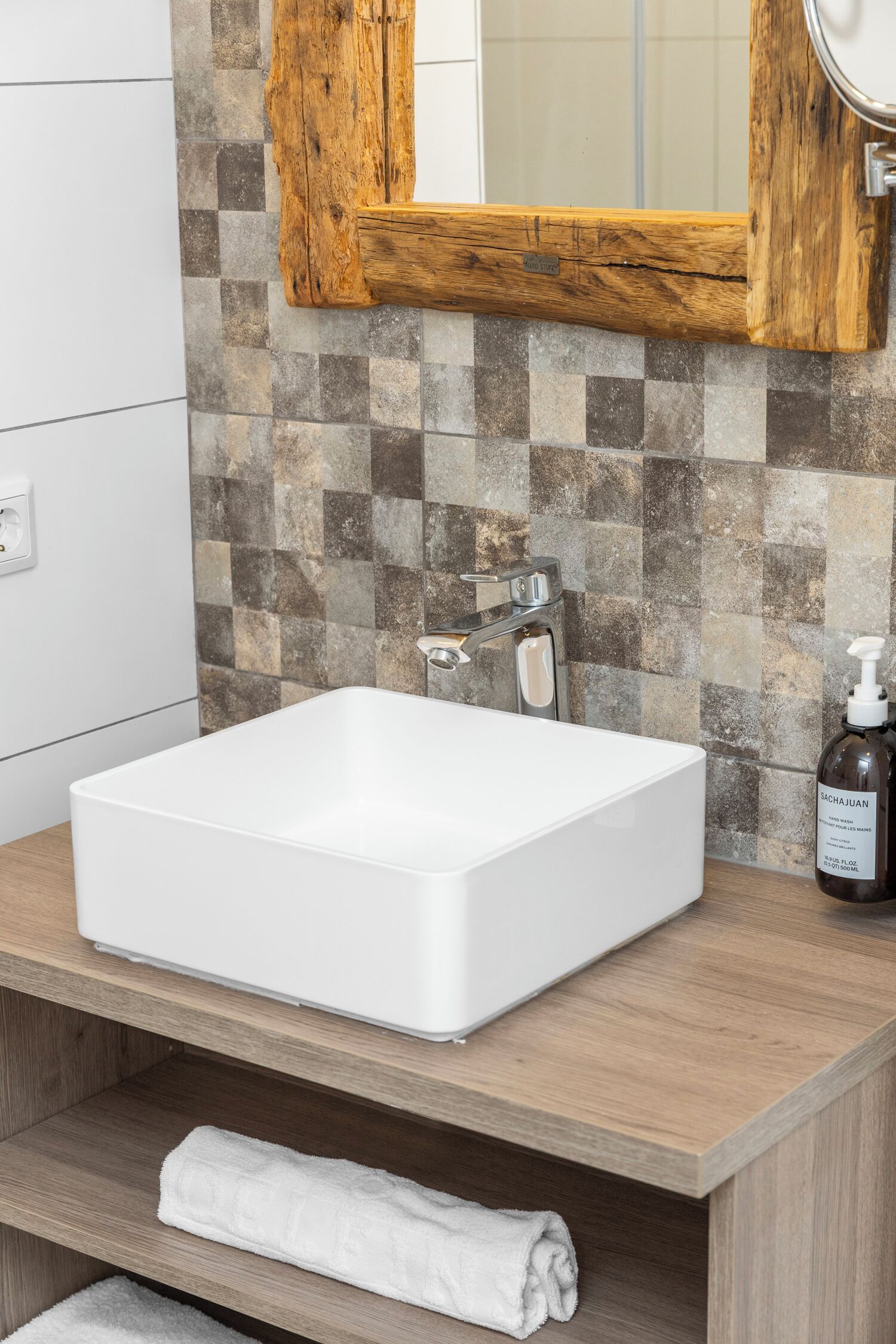 Une salle de bain avec un lavabo blanc, un meuble-lavabo en bois, un miroir et un flacon de savon pour les mains.