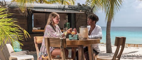 A wooden table with two chairs, a small table lamp, and a menu board in a beachside setting with palm trees and a view of the ocean.