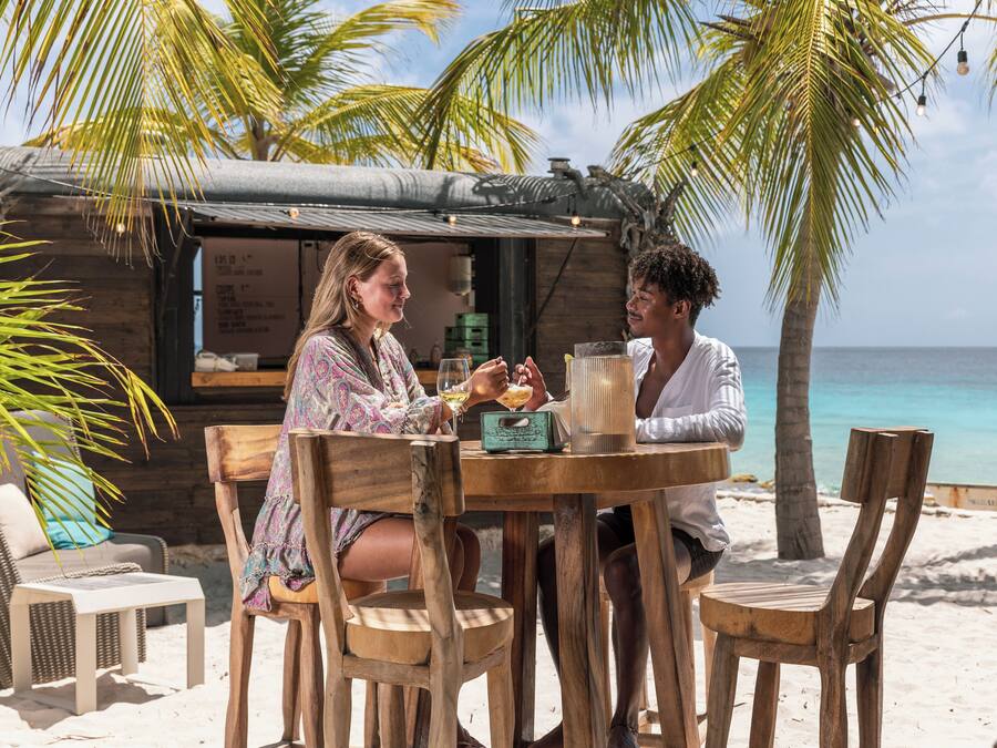 A wooden table with two chairs, a small table lamp, and a menu board in a beachside setting with palm trees and a view of the ocean.