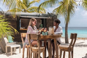 A wooden table with two chairs, a small table lamp, and a menu board in a beachside setting with palm trees and a view of the ocean. - Delfins Beach Resort Bonaire, Tapestry Collection by Hilton (Kralendijk)