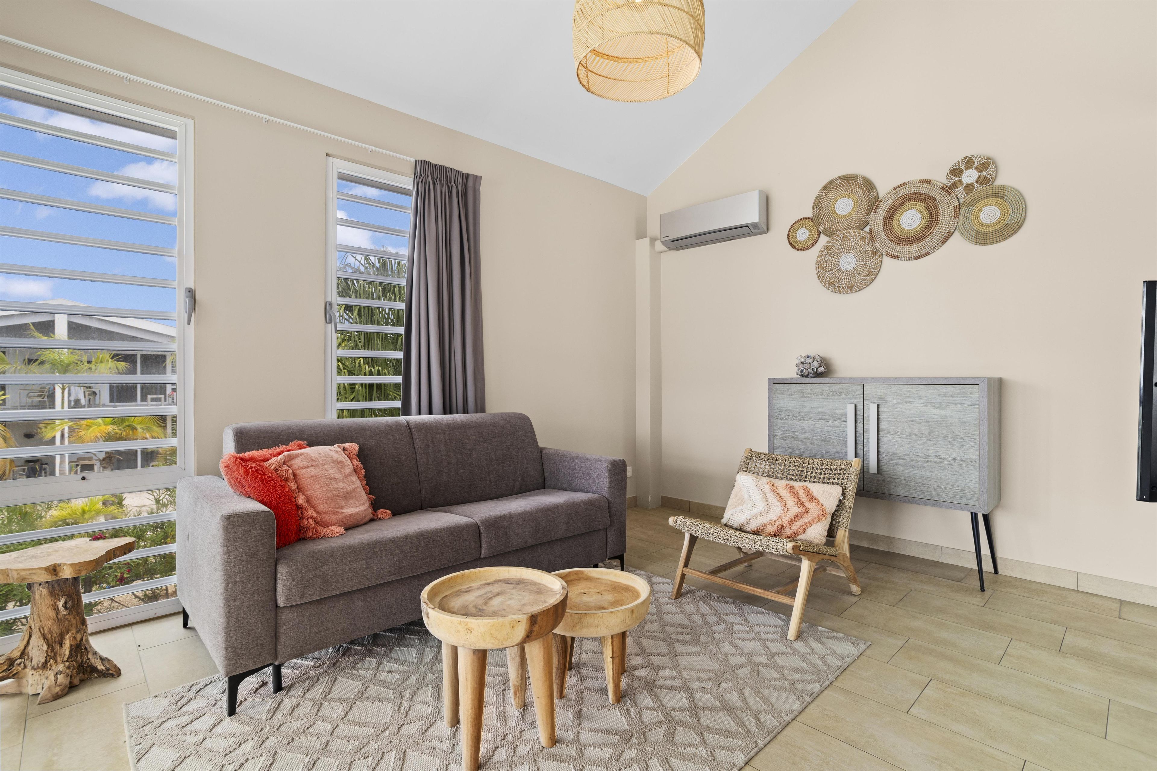 Un salon avec un canapé gris, deux petites tables en bois, un fauteuil en osier, un meuble gris et une grande fenêtre donnant sur un paysage verdoyant.
