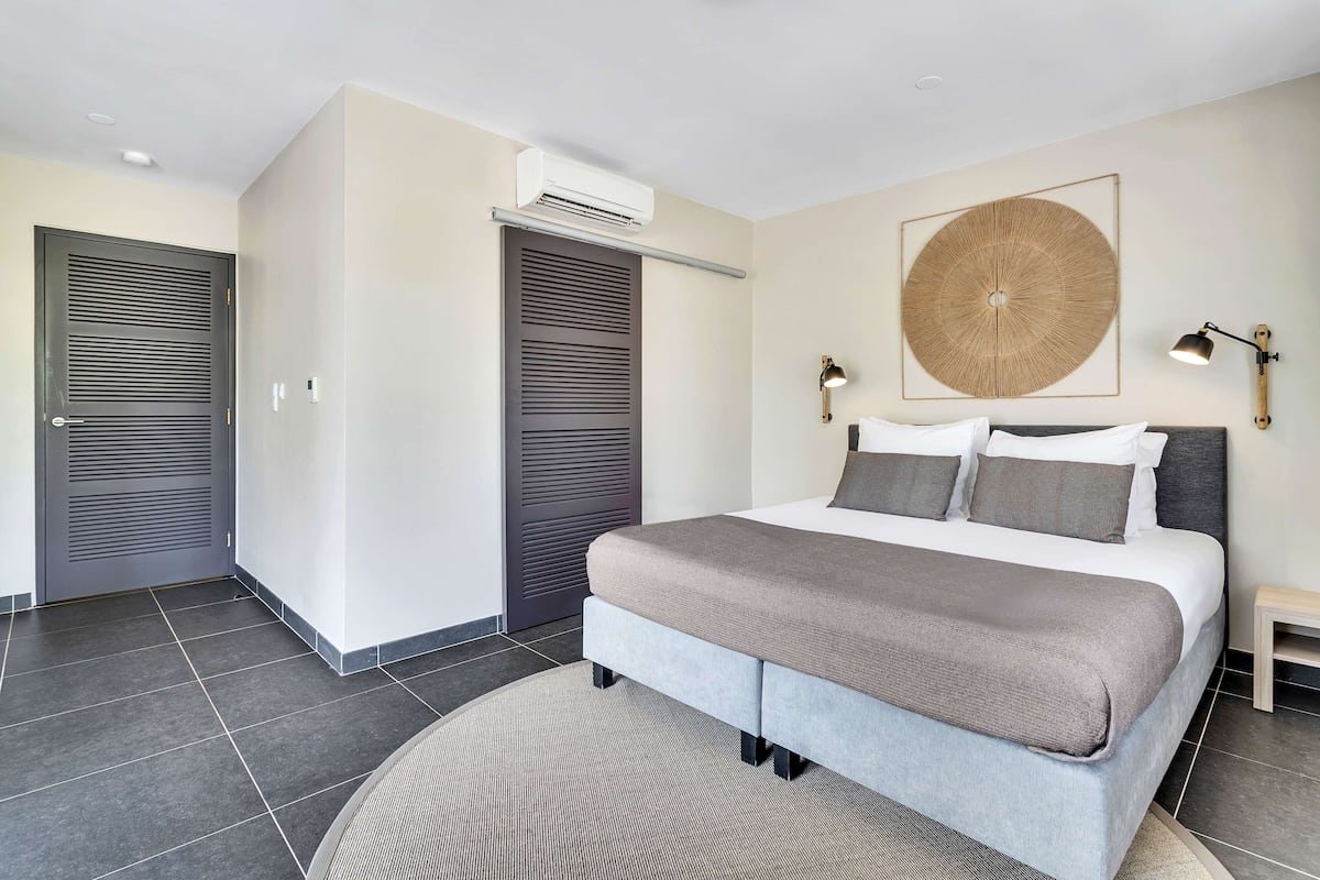 A modern bedroom with a large bed, two bedside tables, an air conditioning unit, and a wall-mounted artwork.