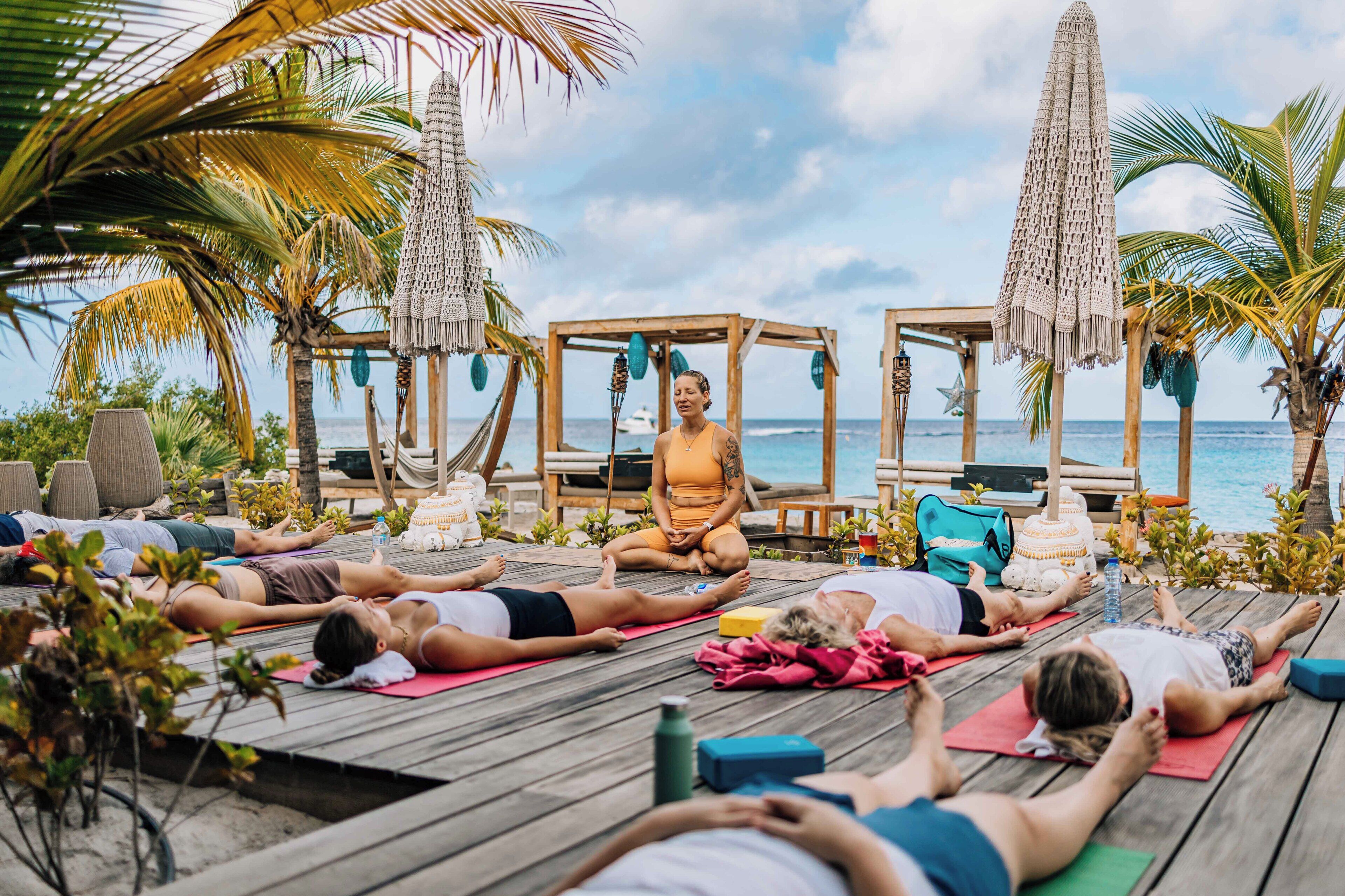 A group of people meditating on a wooden deck by the sea, with palm trees and a clear sky in the background.