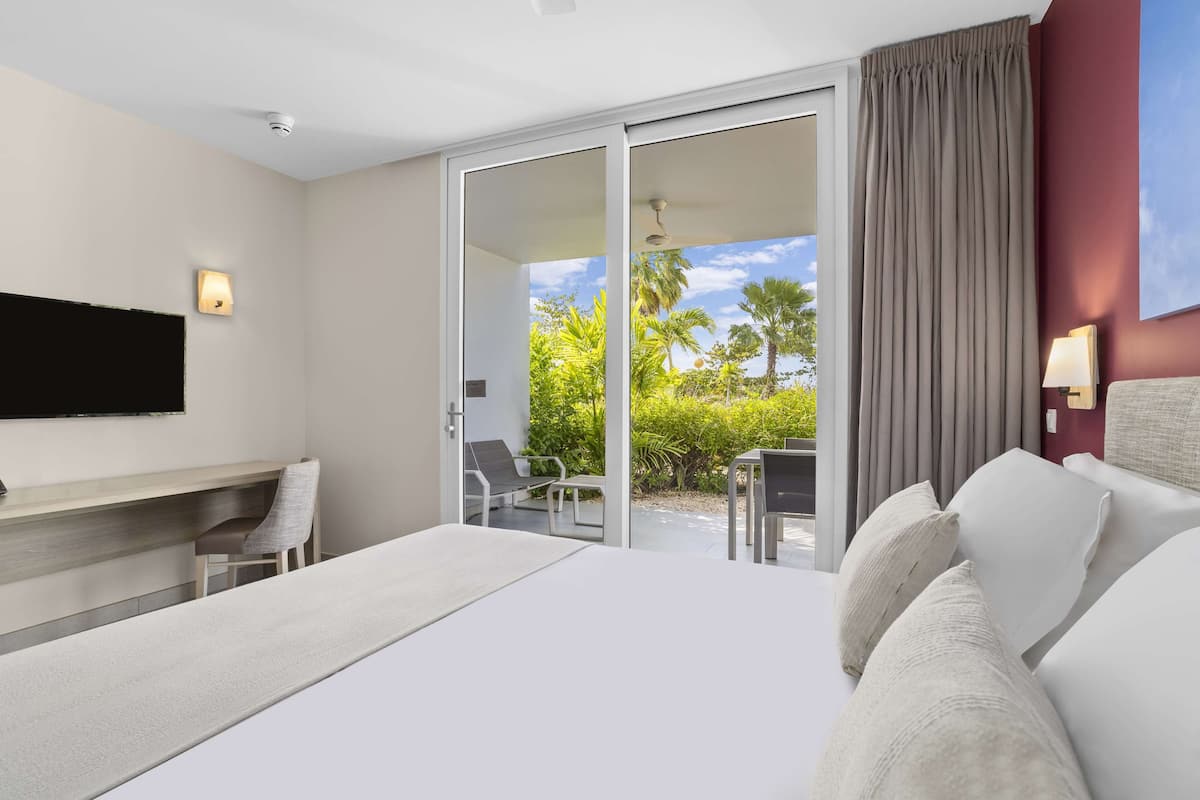 A modern hotel room with a large bed, a flat-screen TV mounted on the wall, a desk with a chair, and a balcony with a view of palm trees and a clear sky.