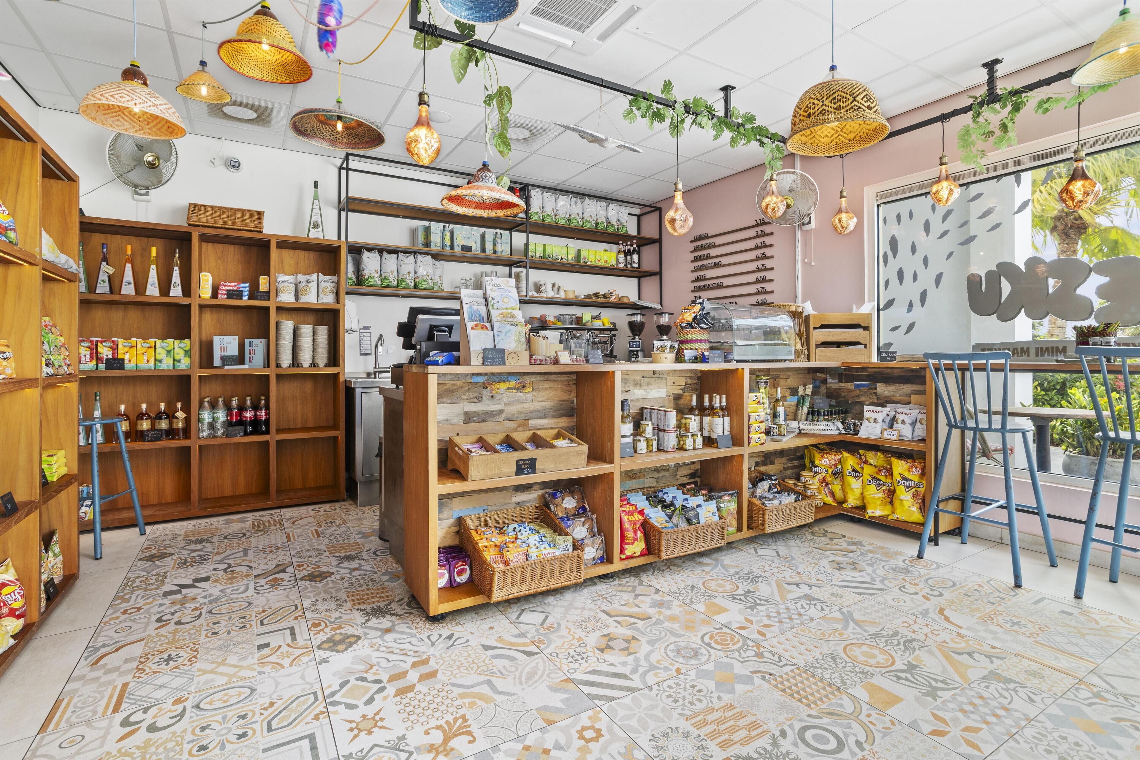 A cozy cafe with wooden shelves stocked with snacks and drinks, a cash register, and hanging light fixtures.